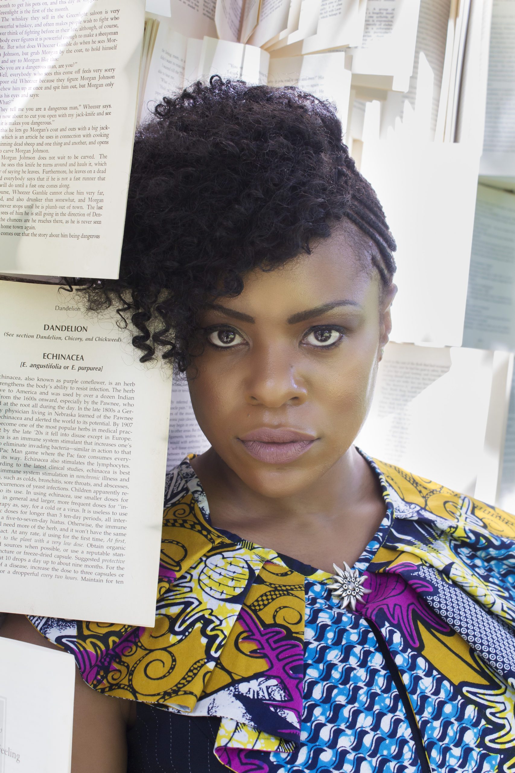 A photo of Kimbilio Fellow, novelist Nana Nkweti.  She wears a colorful blouse and poses surrounded by various manuscripts.  