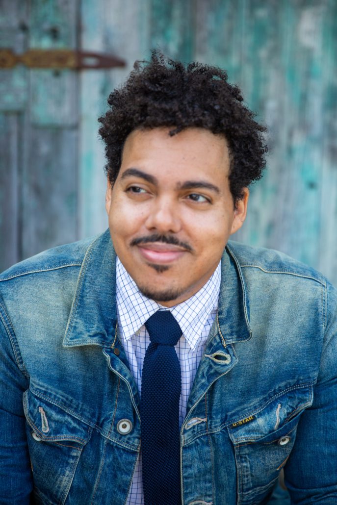 a black man wearing a blue jean jacket sits in front of a blue wall